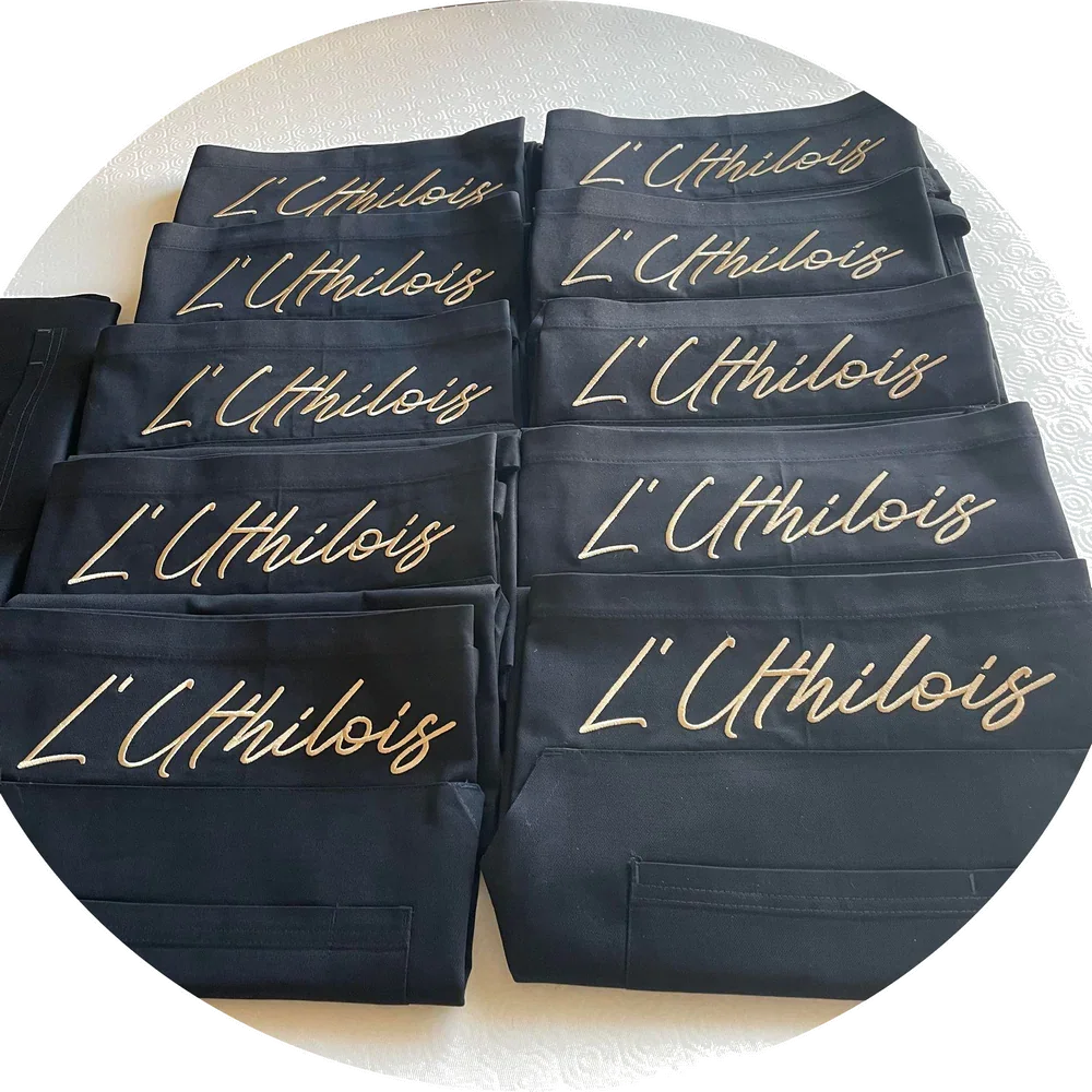 Tabliers noirs brodés "L'Uthilois" en fil doré, empilés sur une nappe blanche, produits personnalisés.
