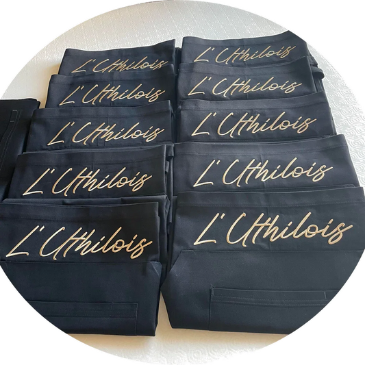 Tabliers noirs brodés "L'Uthilois" en fil doré, empilés sur une nappe blanche, produits personnalisés.