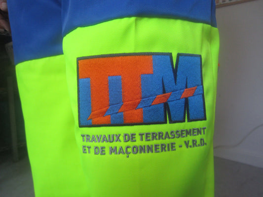 Logo TTM brodé sur tissu jaune fluo, texte travaux de terrassement et maçonnerie, vêtement pro