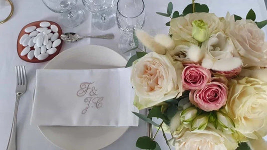 Serviette de table brodée pour mariage, posée sur assiette, bouquet de roses et dragées en déco élégante.