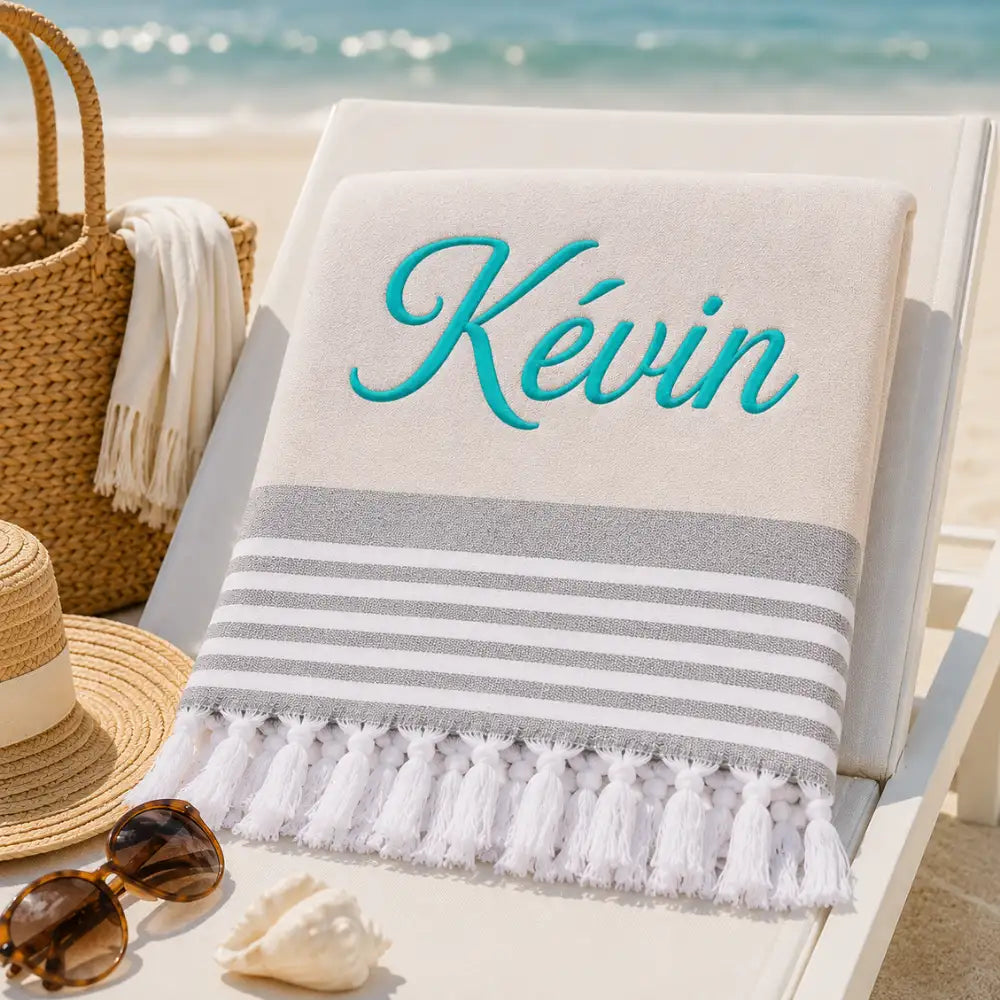 Fouta_personnalisee_beige_naturel_brodee_au_prenom_Kevin_en_fil_turquoise_serviette_plage_coton_turc_a_franges_sur_transat